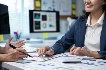 Colleagues discuss financial reports at a desk, reviewing charts on paper and dashboards on monitors with a calculator nearby in a modern office