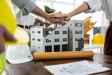 Architects stack hands over a building model with blueprints and hard hats on the table, celebrating teamwork and planning a construction project in a bright studio.