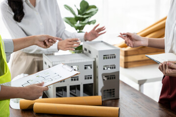 Architects collaborate around a building model, reviewing blueprints and notes during a design meeting with rolled plans and a tablet in a bright office.