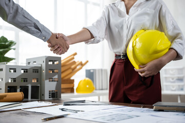 Two construction professionals shake hands over blueprints in an office; one holds a yellow hard hat beside scale building models, signaling a new project agreement.