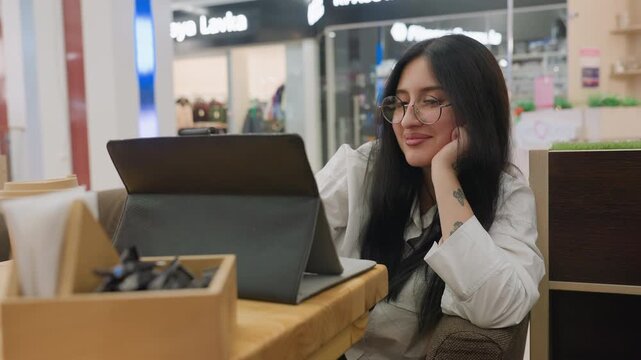 Young woman wearing glasses smiles softly while working on tablet in mall, resting tattooed hand under chin in relaxed position, seated at wooden table with takeaway coffee and utensil box nearby