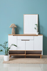 Chest of drawers with lamp, blank frame and plants near green wall in room