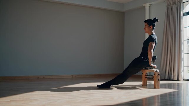 Sportswoman wearing black sportswear performing triceps dips exercise using wooden chair in gym with window and wooden floor during fitness workout