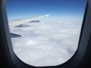Fototapeta premium An expansive aerial view from an aircraft window, showing a vast blue sky stretching over a thick, fluffy blanket of white clouds, with the plane's wing visible
