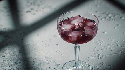 High Angle Shot of Crystal Coupe Glass Filled with Ice and Red Beverage Being Poured on Wet White Surface Capturing Refreshment Elegance and Celebration in Motion with Shadow and Light