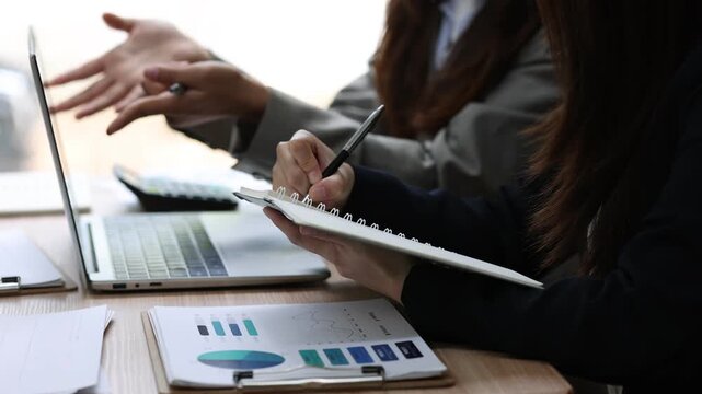 Two businesswoman are sitting at a desk, actively engaged in a collaborative project, utilizing a laptop and writing a memo to analyze data and discuss strategies.