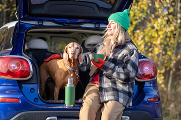 Woman pet owner drinking tea sitting in car trunk with dog enjoying nature in forest on weekend....