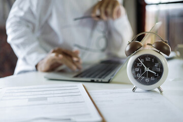selective focus on alarm clock, asian businessman working with laptop and document on table before...