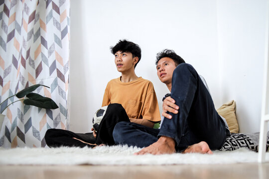 Two Young Asian Men Sitting On Floor While Watching Soccer Match On Television