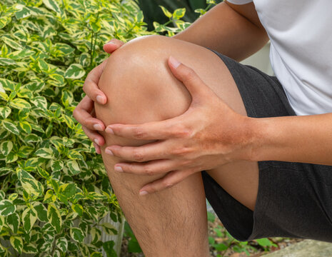 Man clutching knee in outdoor garden—signs of joint pain or ligament strain from sudden movement. Ideal for medical ads on knee injuries, tendon issues, or arthritis.