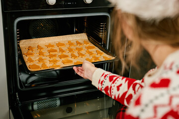 Woman in red festive sweater and Santa hat putting tray of Christmas cookies into oven. Process of cooking traditional gingerbread cookies for winter holidays at home kitchen