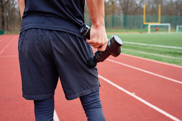Close-up of man recovery muscle after training. Athlete using massage gun on leg while standing on running track. Concept of muscle recovery, sports therapy and injury prevention