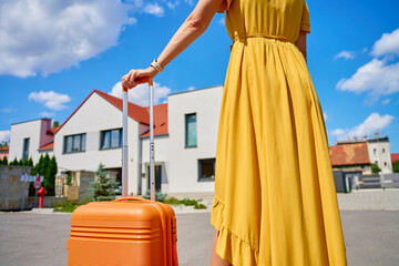 Woman in yellow dress pulls orange suitcase along residential street with houses on sunny day. Female traveler returning home from journey. Concept of travel, homecoming and end of vacation