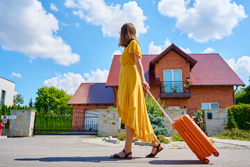 Woman in yellow dress with orange suitcase walking on street near residential house on sunny day. Female traveler with luggage return home from journey