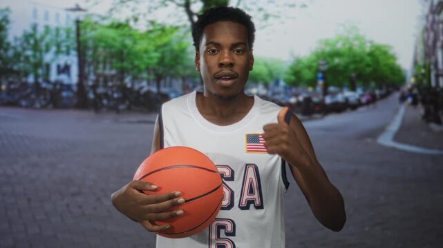 Man holding basketball, smiling and giving thumbs up on street with white jersey and american flag patch; confidence.
