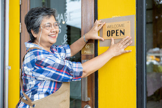 Senior woman hanging open sign on bright yellow cafe door, smiling proudly as she prepares to welcome customers, representing small business ownership and friendly local service.