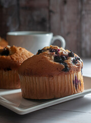 Blueberry bread on a white plate with a blurred coffee cup in the background and a wooden wall....
