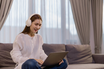 Happy asian female freelancer working remotely from home, using laptop and headphones for video call, sitting comfortably on sofa in living room