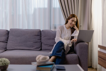 Young woman is sitting on a comfortable sofa in her living room, smiling and working remotely using her laptop, enjoying the convenience and flexibility of working from home