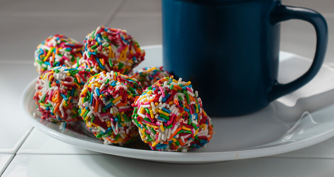 Macro photography of very colorful sweet bread balls for children with a blue cup on one side on a white plate - Powered by Adobe