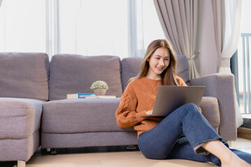 Beautiful asian woman sitting on the floor and using laptop working from home in living room with...
