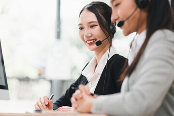 Young woman customer service representative smiling at headset while coworker assists and takes notes in bright office environment showing teamwork and professional support