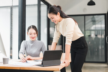Two Asian women collaborating at modern office desk smiling while reviewing documents and tablet...