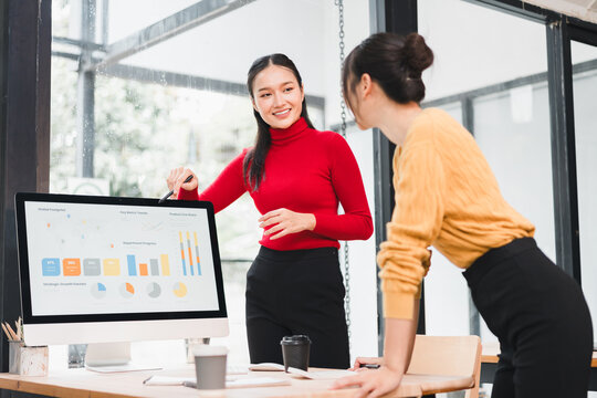 Young woman presenting data on desktop monitor to colleague in modern office, smiling and engaged with colorful charts and graphs, collaborative business meeting with casual professional attire