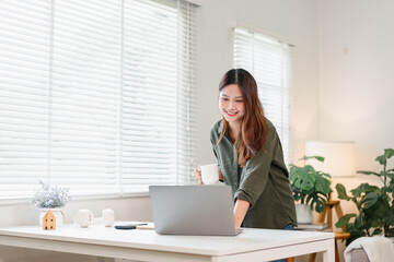 Young woman working on laptop at bright home desk with coffee and plants, smiling and focused in cozy modern workspace