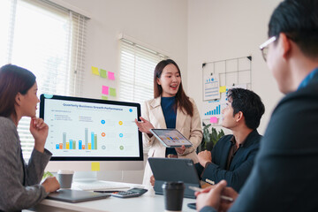 Young businesswoman presenting quarterly growth chart to colleagues in bright meeting room, smiling confidently while pointing at tablet and monitor, collaborative professional teamwork atmosphere
