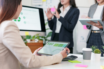 Young businesswoman holding tablet displaying ESG dashboard while colleagues present strategy in modern office with plants and coffee