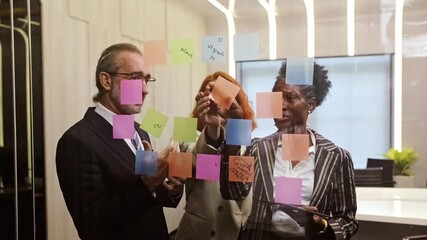 Three colleagues brainstorm in a modern office, placing colorful sticky notes on a glass wall during a planning meeting, highlighting teamwork, creativity, and collaborative strategy. 4k video