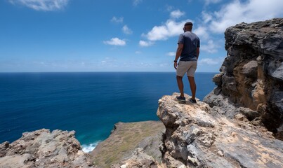 a man stands on top of the rocky cliff, looking down at us from above, with his back to the viewer. the sky is blue and clear, and there are some white clouds in it