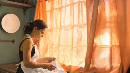 Fototapeta premium A thoughtful woman in a simple apron meticulously folds white linen, illuminated by warm, golden light streaming through an orange-draped window.