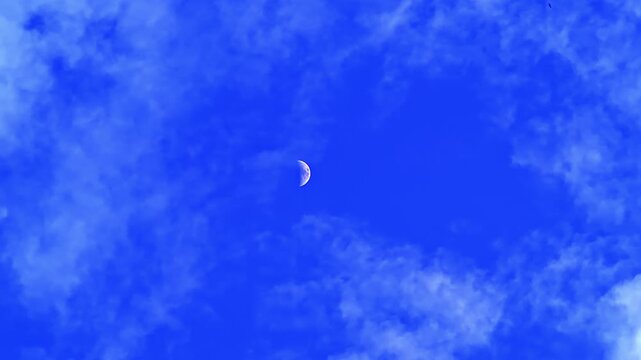 4K footage of a crescent moon in a bright blue sky with moving clouds during daytime, creating a serene and natural atmosphere.