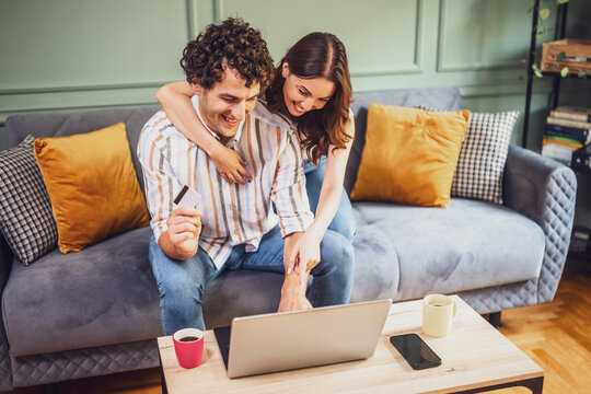 A smiling couple uses a laptop to pay for online shopping, enjoying a fun and relaxed moment together in the living room.
