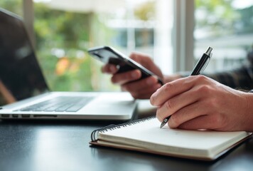 Person Writing in Notebook at Home Office Desk with Laptop and Smartphone