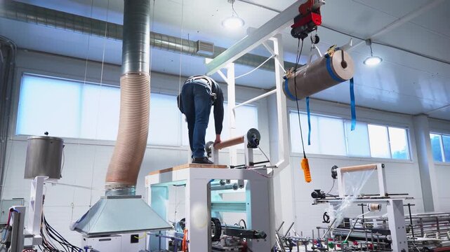 Worker climbs onto production line chamber to repair issue in workshop backside view. Technician checks extrusion machine tool at manufacturing plant