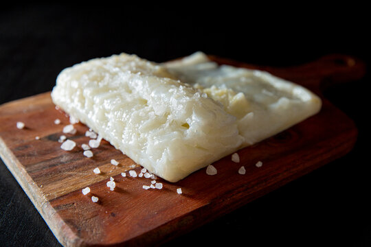 Raw lutefisk on wooden board with coarse salt, dramatic studio light on black background, minimalistic food concept.