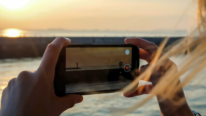 Woman shooting video of lighhouse on smartphone at sunset. Close up