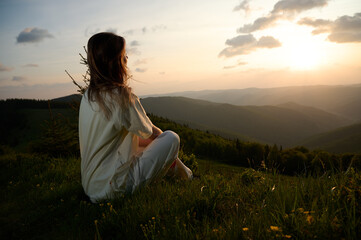 Woman sits peacefully on a grassy hill, gazing at the sunset as golden light bathes the landscape....