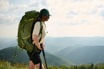 Woman hiker stands on a grassy ridge, taking in the vast mountain scenery. Dressed in a cap and...