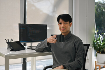 Young Asian male programmer smiles confidently while holding a black coffee mug in a modern office, with code visible on screen