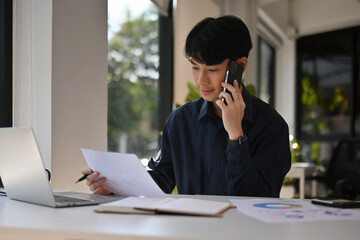 Young Professional on Business Call, Businessman Talking on Phone While Reviewing Reports at Desk