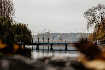 An autumn scene shows a long, pedestrian bridge over a calm lake under a soft, overcast sky. The...