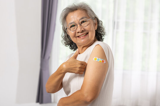 Asian Elderly woman smiling with thumb up showing colorful adhesive bandage on upper arm after vaccination indoors