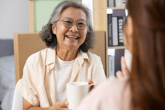 Asian Elderly woman smiling and holding white mug while having conversation with another person in cozy indoor setting, expressing warmth and happiness