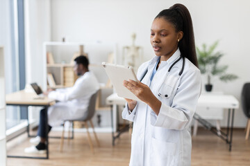 Female adult healthcare professional dressed in white coat using digital tablet in office setting. Office environment includes another healthcare worker seated in background working on computer.