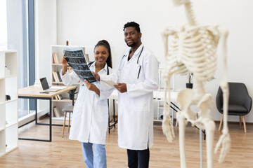 Male and female doctors wearing lab coats examining X-rays in medical clinic. Skeleton model...