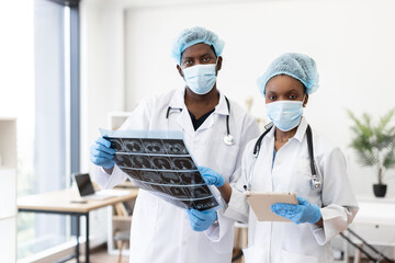 Two African American surgeons wearing masks in medical consultation, reviewing patient scans, collaborating in hospital. Their focus emphasizes teamwork and healthcare expertise